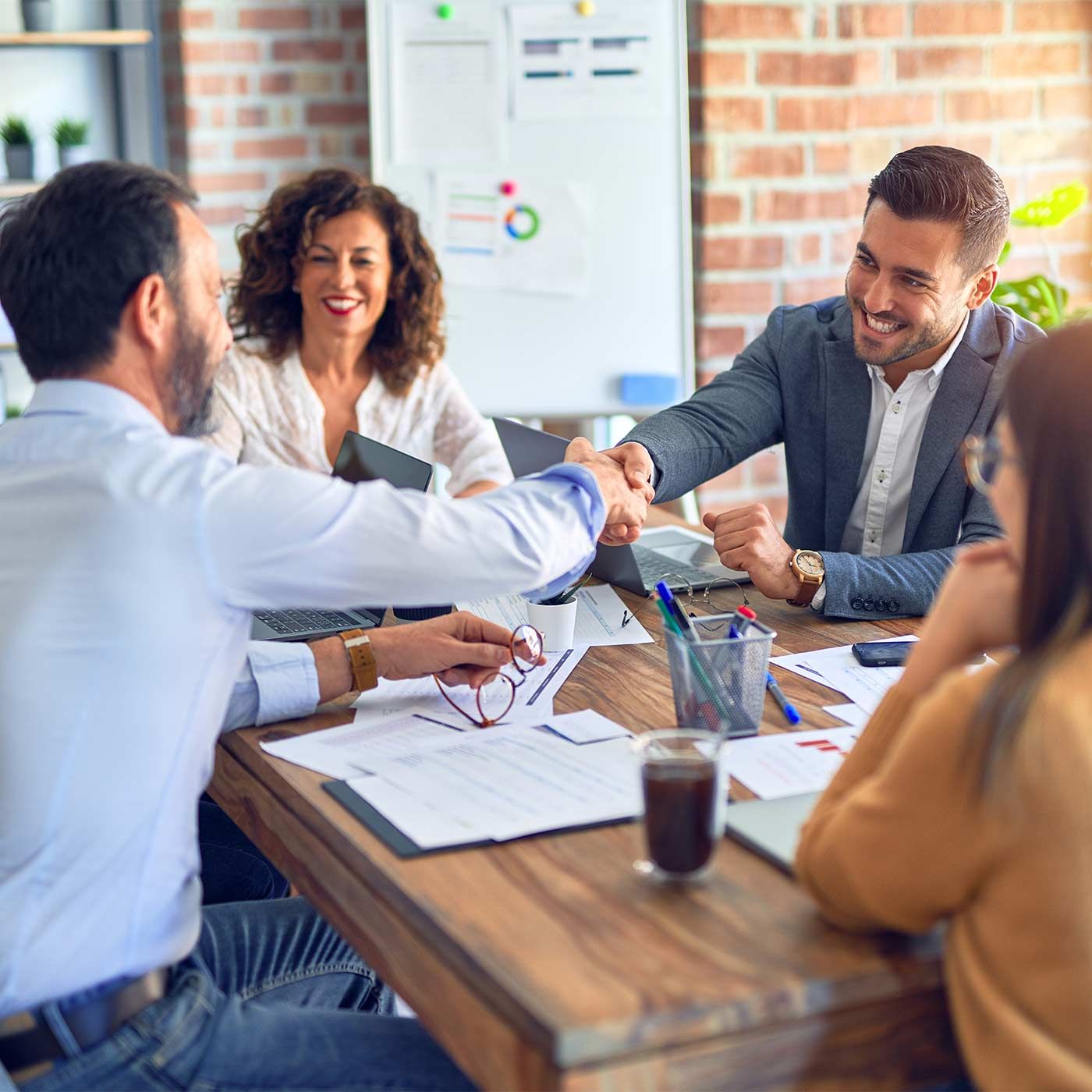 Twee zakenmannen schudden elkaar de hand terwijl collega's toekijken en glimlachen.